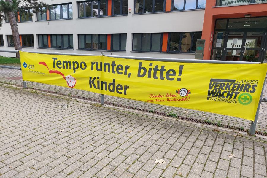 Blich auf eine Schule. An einem Geländer hängt ein großes gelbes Plakat mit der Aufschrift: Tempo runter, bitte! Kinder