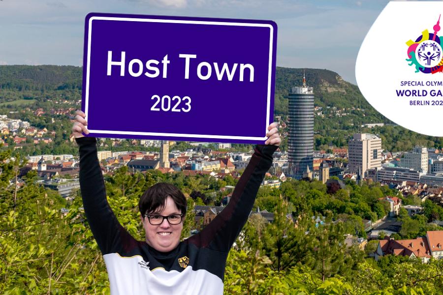Frau in Sportsachen hält Jena-Schild hoch vor Stadtkulisse