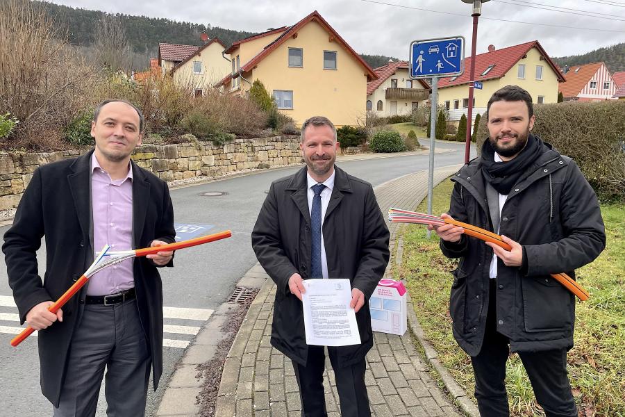 Drei Männer scheuen in die Kamera, im Hintergrund eine Siedlung mit Einfamilienhäusern