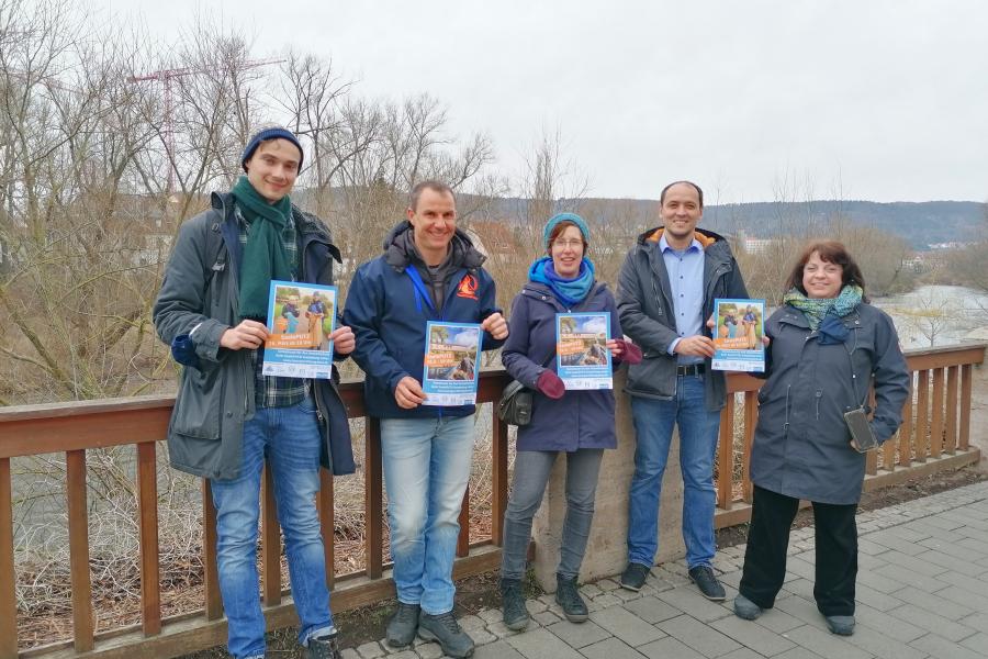 5 Menschen halten Plakate in die Kamera, hinter ihnen ein Fluss