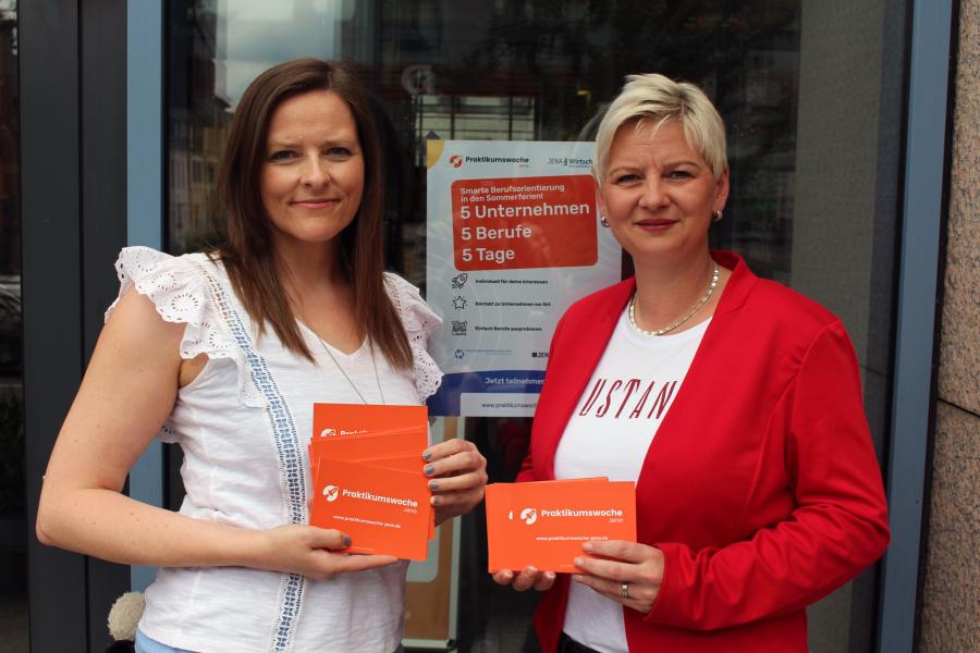 Zwei Frau stehen vor einer Glastür und schauen in die Kamera. Sie halten Flyer in der Hand. 