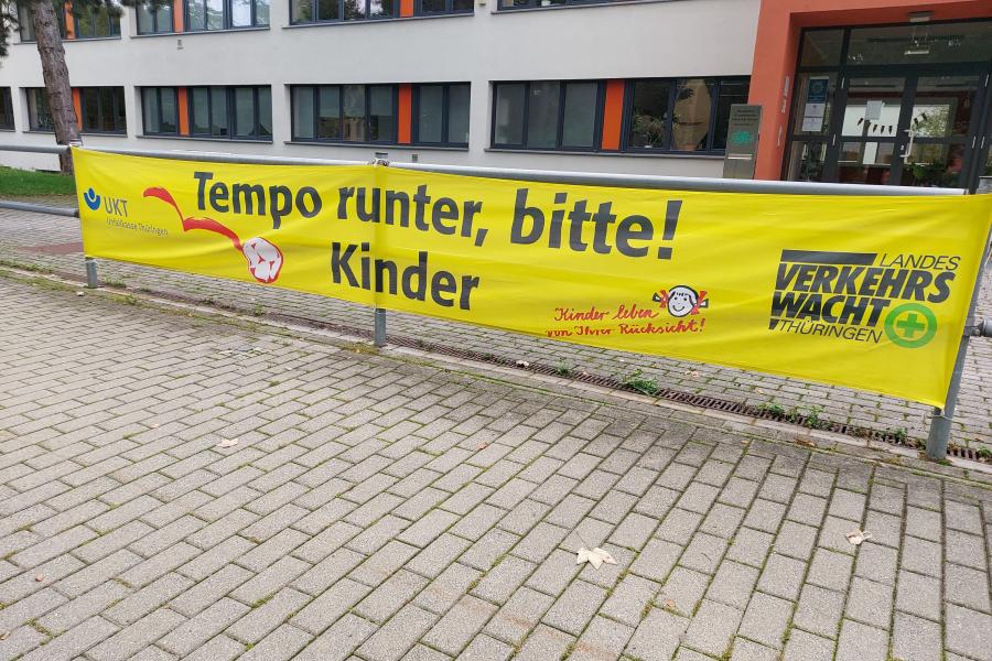 Vor einer Schule hängt ein Banner, dass zu Vorsicht im Verkehr aufruft.