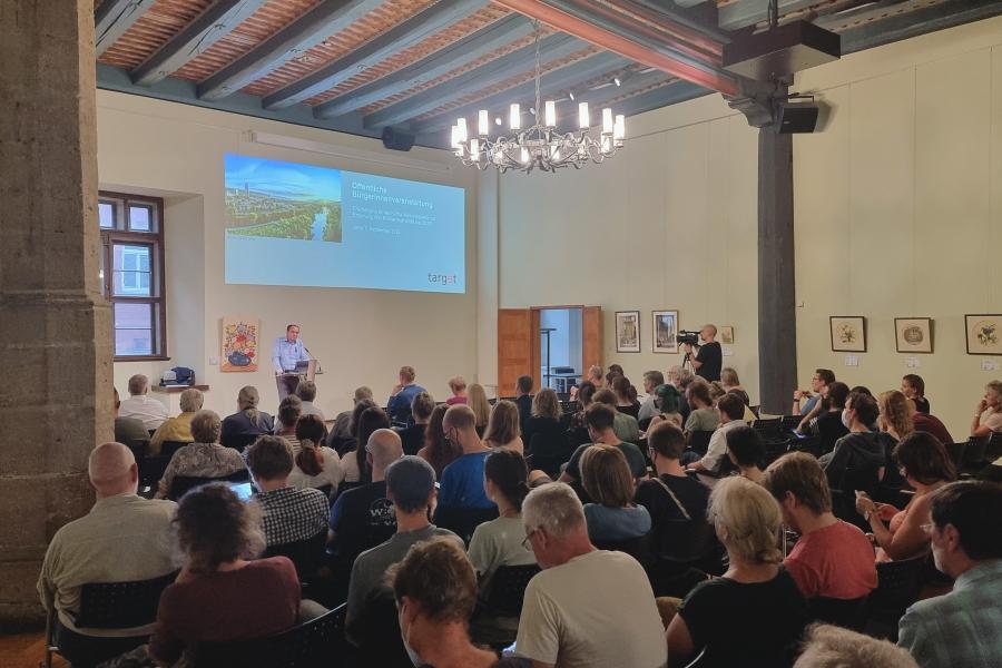 Bürgermeister Christian Gerlitz steht auf einer Bühne am Pult und begrüßt viele Menschen im Historischen Rathaus.
