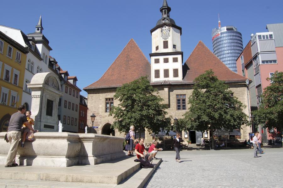 Im Vordergrund ein steinerner Brunnen auf einem großen Platz, im Hintergund mehrere Gebäude.