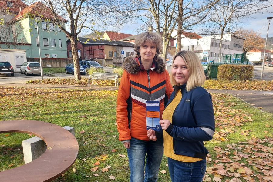  Dörthe Thiele (l.) und Kathrin Bajohr vom Büro für Migration und Integration stehen auf der Wiese vor dem Verwaltungsgebäude am Anger 15 und halten einen Flyer der Antidiskriminierungsstelle in der Hand.