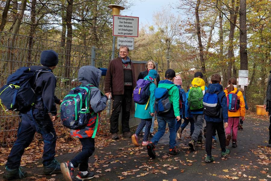 Grundschulkinder gehen durch ein geöffnetes Tor. 3 Erwachsene empfangen Sie.