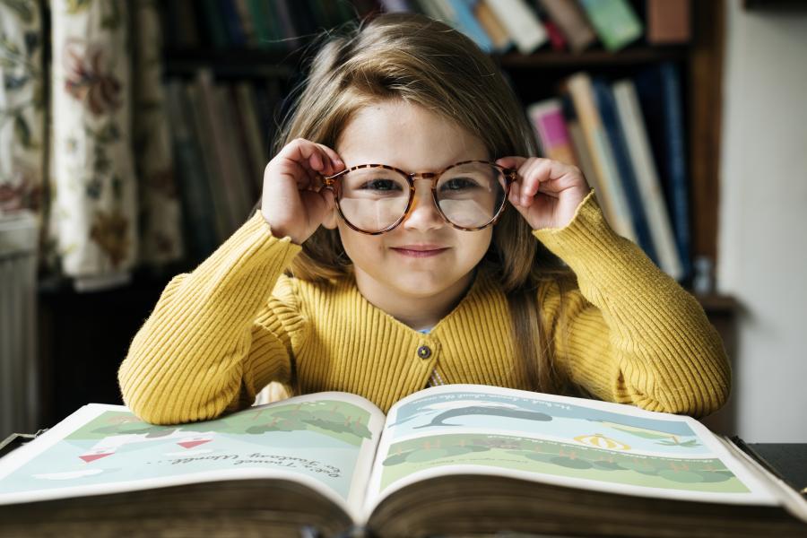 Ein Mädchen mit Brille sitzt an einem Tisch, vor ihr liegt ein großes, aufgeschlagenes Buch.