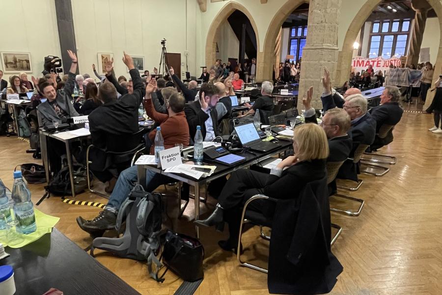 Abstimmung des Jenaer Stadtrats: Die meisten Stadträte heben die Hand. Im Hintergrund halten Personen Transparente hoch und fordern den Klima-Aktionsplan zu beschließen.