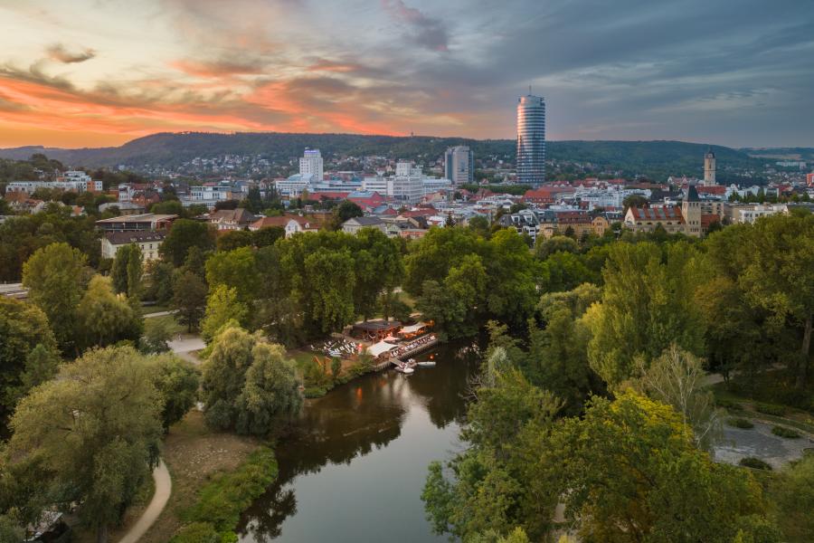 Panoramaaufnahme der Stadt Jena mit einem Fluss und viel Grün runsherum im Vordergrund und Bebauung im Hintergrund