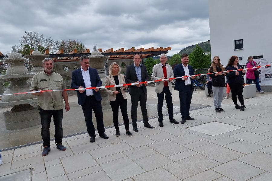 Mehrere Personen schneiden ein Band vor einem Platzmit Brunnen durch