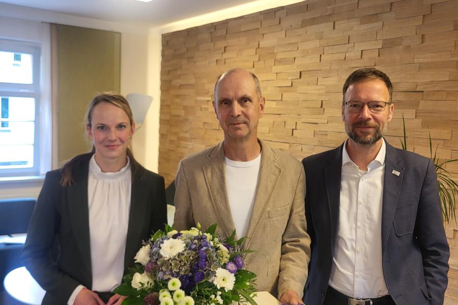 3 Personen stehen in einem Raum, eine Person hält Blumen und eine Urkunde in der Hand.