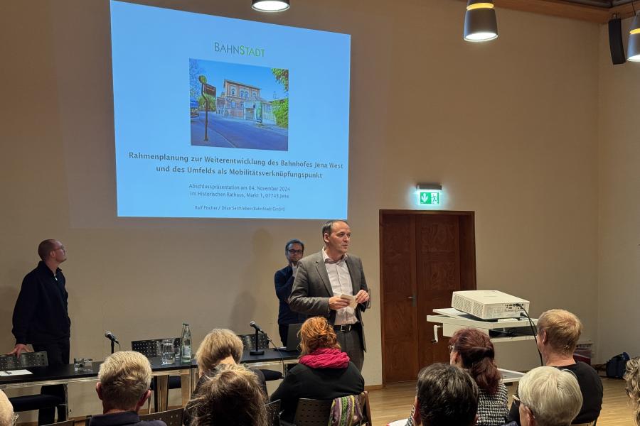 Bürgermeister Christian Gerlitz stehtim Plenarsaal im Historischen Rathaus und spreicht vor sitzenden Personen. HInter ihm ist eine Präsentation via Beamer an die Wand geworfen zum Thema Rahmenplanung zur Weiterentwicklung des Bahnhofes Jena West