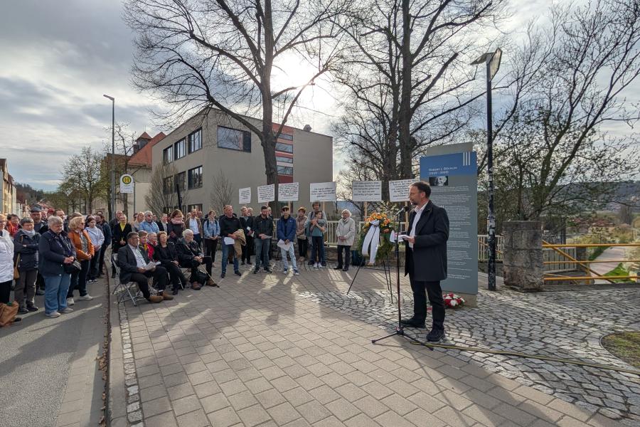 An einer Gedenkstele auf dem Gehweg vor dem Angergymnasium steht OB Thomas NItzsche und hält an einem Mikrofon eine Rede. Vor der Stele liegen Blumen, damit steht ein Kranz auf einem Ständer. Im Halbkreis um die Stele stehen Menschen unterschiedlichen Alters und höheren zu, einige sitzen auf Stühlen.