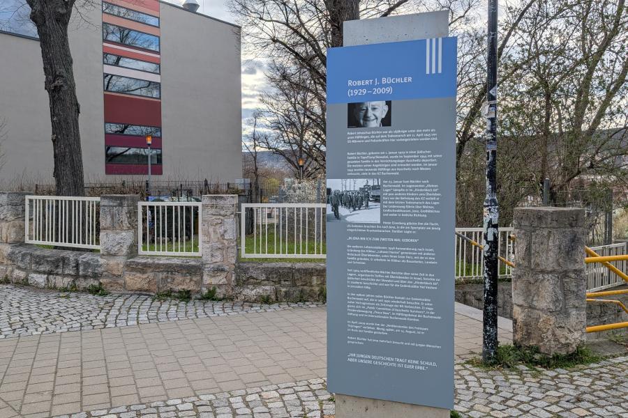 Eine Stele mit Informationen zu Robert J. Büchler und dem Todesmarsch am 11. April 1945 steht auf einem Gehweg. Im Hintergrund ist ein Schulgebäude zu sehen.