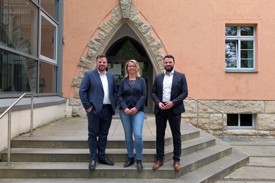 Zwei Männer und eine Frau stehen nebeneinander auf der Treppe, die zum Haupteingangs des technihschen Rathauses in Jena führt.