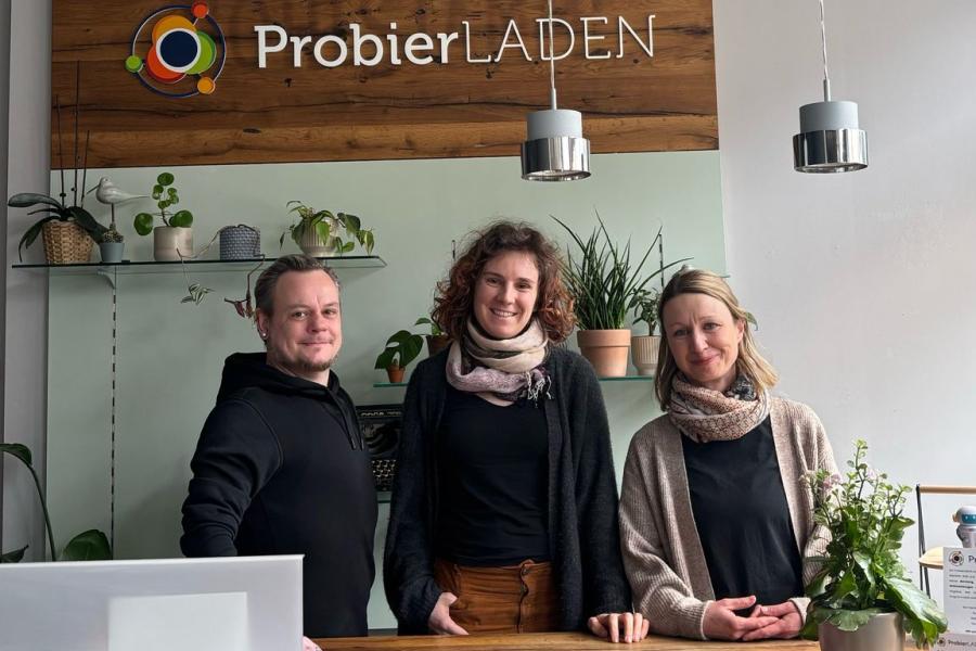 Ein Mann und zwei Frauen stehen hinter einer Empfangstheke. Dahinter hängt ein Bild mit der Aufschrift ProbierLaden.