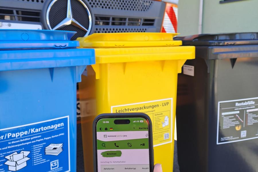 Handy mit abgebildeten Behälter-Konfigurator vor einer blaueb, gelben und scharzen Mülltonne sowie einem Müllfahrzeug