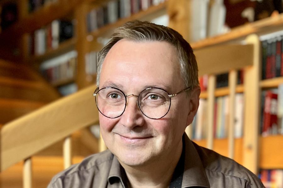 Ein Mann um die 50 sitzt vor einer Holztreppe, trägt eine Brille und ein hellbraunes Hemd.