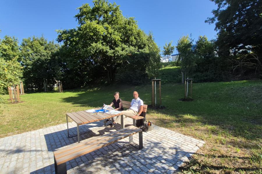 Naturnahe Lichtung mit Rastplatz: Eine Frau und ein Mann sitzen in der Sonne auf einer Bank.