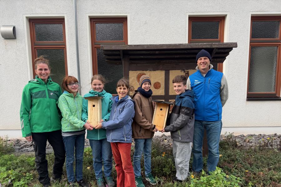 Fünf Jugendliche (drei Mädchen und zwei Jungen) halten zwei Nistkästen aus Holz. Links neben ihnen steht eine Frau und auf der rechte Seite ein Mann.