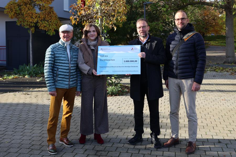 v.l.: Friedrich Wilhelm Gebhardt, ehemaliger Ortsteilbürgermeister von Jena-Winzerla, Ostbeauftragte Elisabeth Kaiser, Oberbürgermeister Thomas Nitzsche und Lars Liebe, Fachdienstleiter Stadtentwicklung stehen nebeneinander und halten einen großen Scheck.