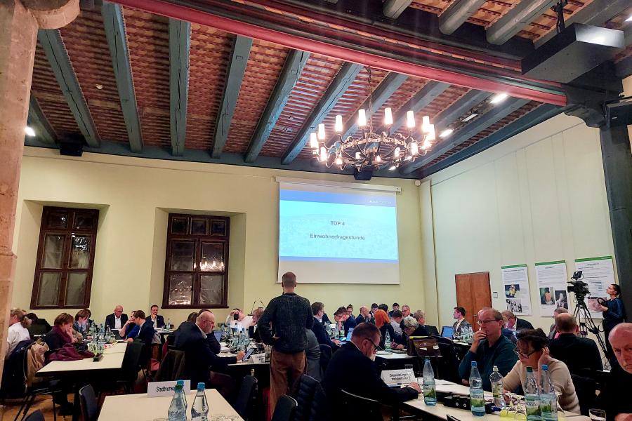 Stadtratsmitglieder sitzen an Tischen im historischen Rathaus, ein Redner steht am Mikrofon