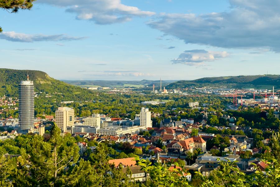 Das Foto zeigt eine weite Panoramaaufnahme der Stadt Jena in Thüringen, aufgenommen vom Landgrafenblick aus. Im Vordergrund sind grüne Baumkronen zu sehen, dahinter breitet sich die Stadt mit ihren Wohnhäusern und Bürogebäuden aus. Links ragt der runde Jentower deutlich über die übrigen Gebäude hinaus. Im Hintergrund ziehen sich die bewaldeten Hügel des Saaletals entlang, und der Himmel ist leicht bewölkt mit Sonnenlicht, das die Landschaft in warmes, freundliches Licht taucht.
