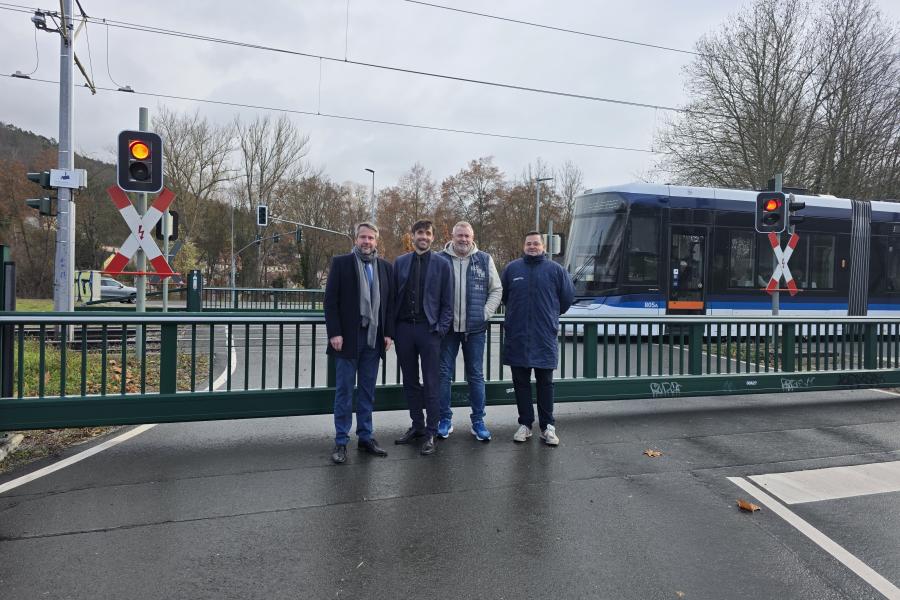 Vier Männer stehen auf der Straße vor einer geschlossenen Schranke. Im Hintergrund fährt eine Straßenbahn vorbei. 