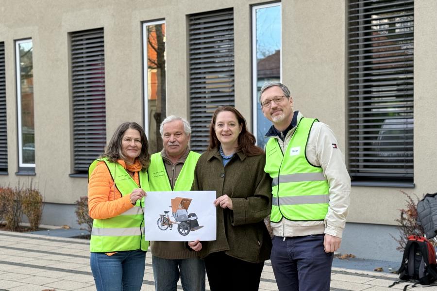 Vier Personen stehen nebeneinander vor einem modernen Gebäude mit großen Fenstern. Drei von ihnen tragen leuchtend gelbe Warnwesten, eine Person in der Mitte trägt eine dunkelgrüne Jacke. Die beiden Personen links halten gemeinsam ein Foto einer Rikscha, die für das Projekt »Radeln ohne Alter« vorgesehen ist. Alle lächeln in die Kamera.