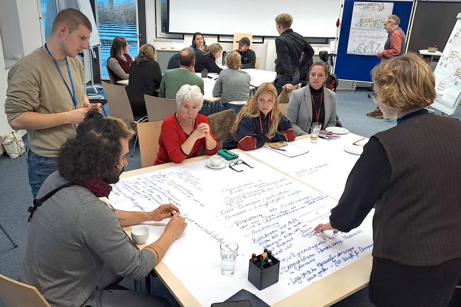 In einem hellen Seminarraum sitzen mehrere Gruppen von Menschen an Tischen und arbeiten gemeinsam an großen Papierbögen. Im Vordergrund beugen sich Teilnehmende über ein großflächiges Blatt, auf dem viele handgeschriebene Stichpunkte stehen. Eine Frau zeigt auf einzelne Notizen, während andere aufmerksam zuhören. Im Hintergrund sind weitere Kleingruppen im Gespräch, Moderationsmaterialien und beschriebene Flipcharts sichtbar. Die Stimmung wirkt konzentriert und kooperativ.