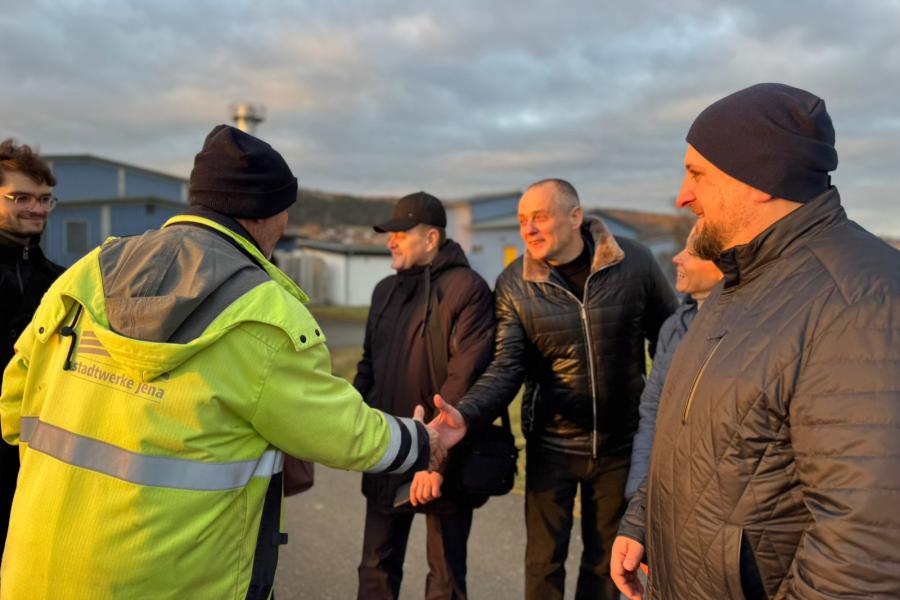 Eine Person in einer gelben Jacke der Stadtwerke Jena gibt mehreren Besuchern im Freien die Hand. Die Gruppe steht lächelnd beisammen, im Hintergrund sind Gebäude und ein bewölkter Himmel zu sehen, das Licht der tiefstehenden Sonne fällt warm auf die Szene.