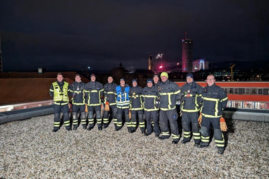 Mehrere Feuerwehrmänner schauen in die Kamera, im Hintergrund Feuerwerk.