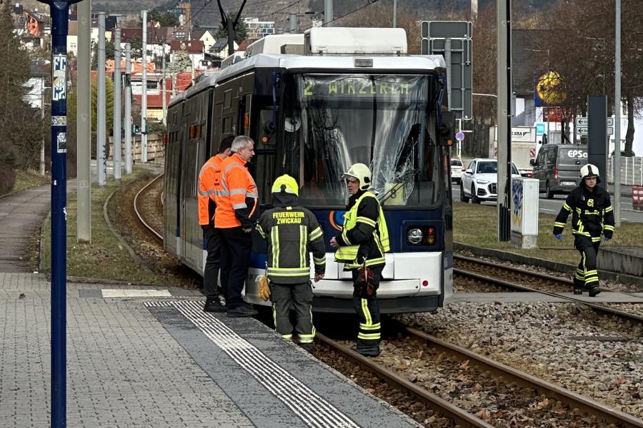 Eine Straßenbahn steht auf den Gleisen mit gesprungener Frontscheibe. Drei Feierwehrleute und zwei Männer in orangfarbener Weste begutachten den Schaden.