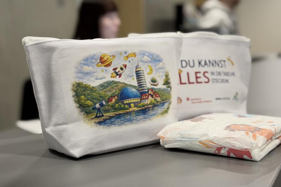 Zwei weiße Stofftaschen mit buntem Aufdruck stehen auf einem Tisch, daneben liegen Windeln; im Hintergrund unscharf Personen in einem Innenraum.