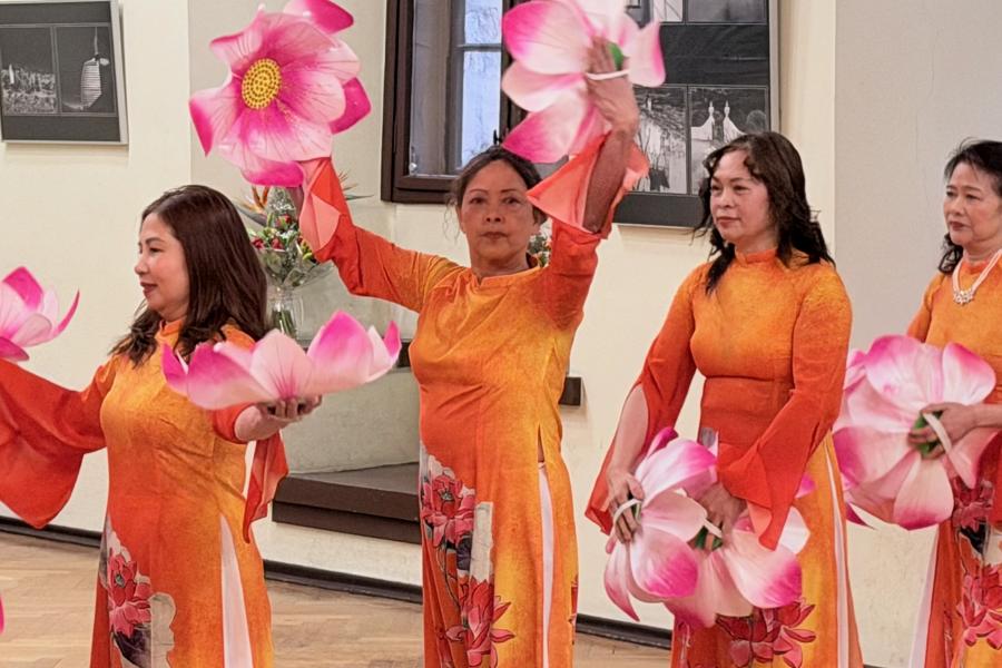 Mehrere Frauen in orangefarbenen traditionellen Kleidern führen einen Tanz mit großen rosa Lotusblüten in einem hellen Ausstellungsraum auf.