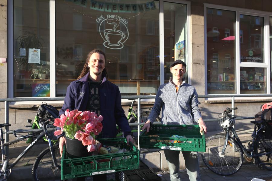 Zwei Männer stehen vor einem Geschäft und halten je eine große Klappkiste mit Lebensmittel und Blumen in der Hand