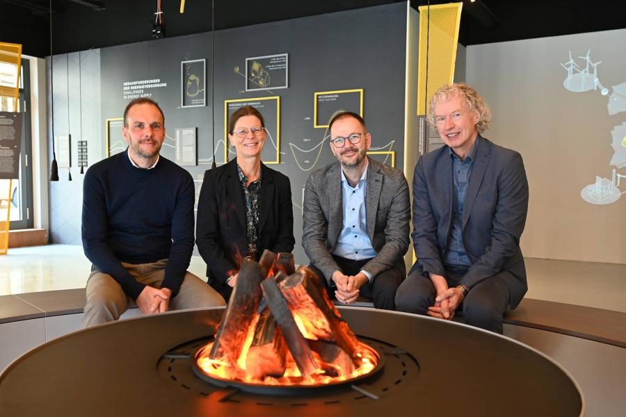 Drei Männer und eine Frau sitzen an einem künstlichen Lagerfeuer in einer Ausstellung.