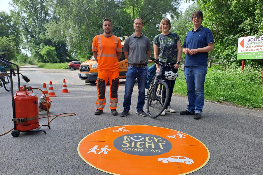 Drei Männer und eine Frau mit Fahrrad stehen hinter einer runden Markierung auf der Straße.