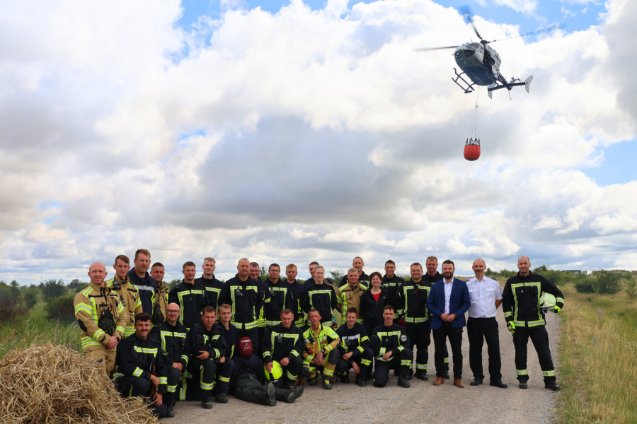 Mehrere Männer stehen auf einem weiten Wiesenfeld, ein Hubschrauber im Hintergrund.