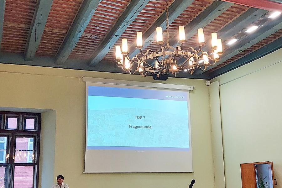 Im historischen Rathaus steht eine Person an einem Rednerpult und andere Menschen sitzen an Tischen, im Hintergrund ist eine Präsentation an die Wand projiziert, darauf steht das Wort "Fragestunde"
