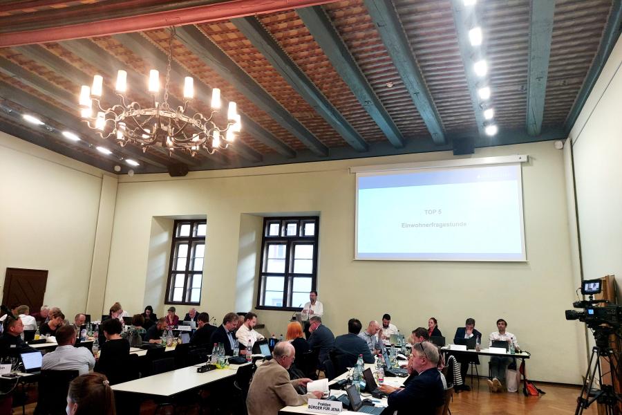 Menschen sitzen an Tischen im Historischen Rathaus, ein Mann steht an einem Rednerpult, im Hintergrund ist eine Präsentation an die Wand geworfen.