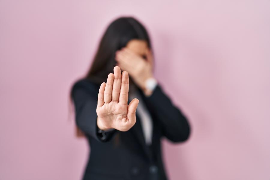 Eine dunkelhaarige Frau hält sich die eine Hand vors Gesicht, mit der anderen macht sie eineStopp-Geste
