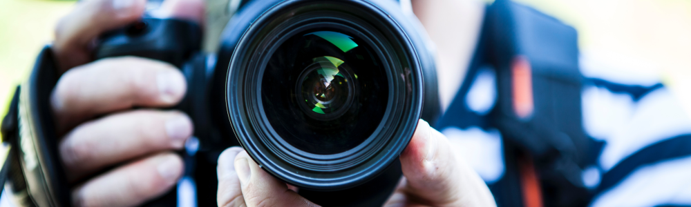 Ein Mann hält eine Fotokamera in der Hand, der Betrachter des Bildes sieht direkt in das Objektiv